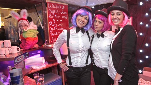 Drei Frauen mit Perücken und Hüten stehen an einem Stand mit Getränken und Speisen.