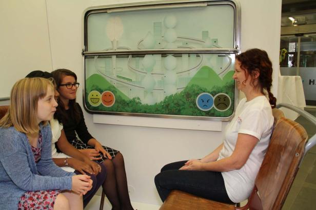 Drei Mädchen sitzen vor einer Frau und einer Darstellung von Emotionen.