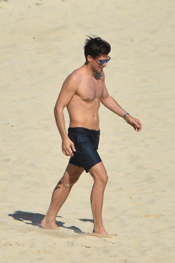 Ein Mann mit Sonnenbrille und Badehose geht barfuß am Strand entlang.