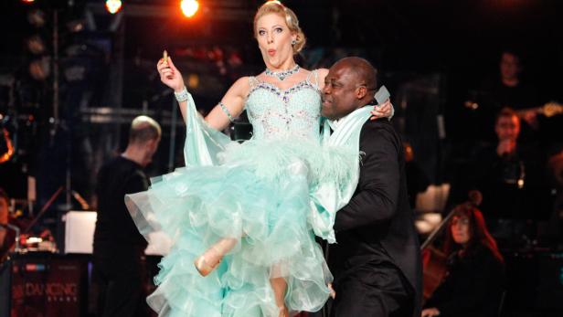 Ein Mann im Anzug trägt eine Frau im Ballkleid auf einer Bühne.