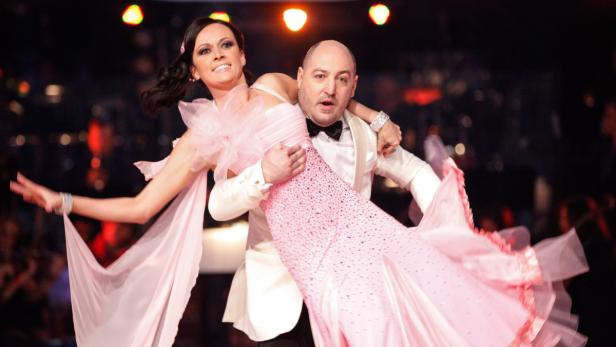 Ein Mann im Smoking trägt eine Frau im rosa Ballkleid auf einer Bühne.