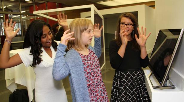 Drei junge Frauen stehen vor einem Bildschirm und halten ihre Hände hoch.