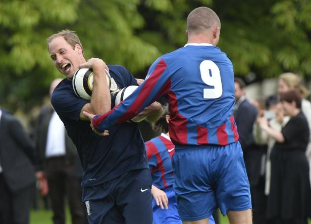 Prinz William wird von einem Fußballspieler mit der Nummer 9 bedrängt, während er mehrere Fußbälle hält.