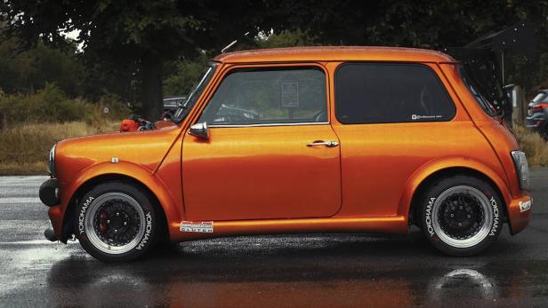 Ein orangefarbener Mini Cooper mit einem großen Heckflügel steht auf einer nassen Straße.