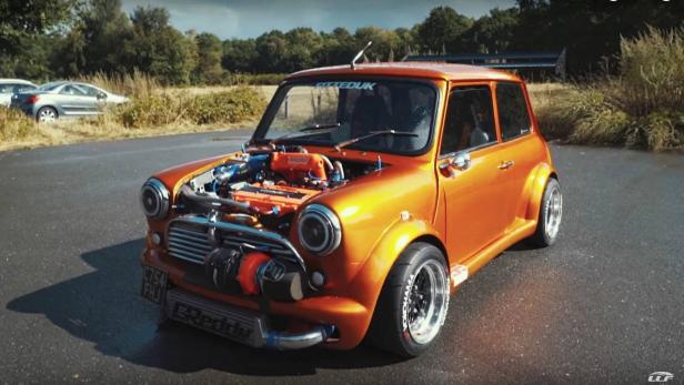 Ein orangefarbener Mini Cooper mit geöffneter Motorhaube und modifiziertem Motor steht auf einer Straße.