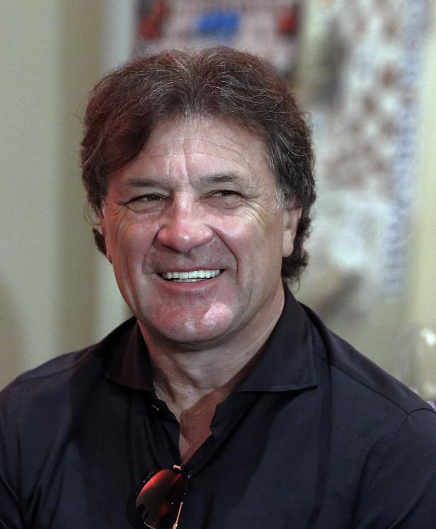 Ein lächelnder Mann mit dunklem Hemd und Sonnenbrille vor unscharfem Hintergrund.