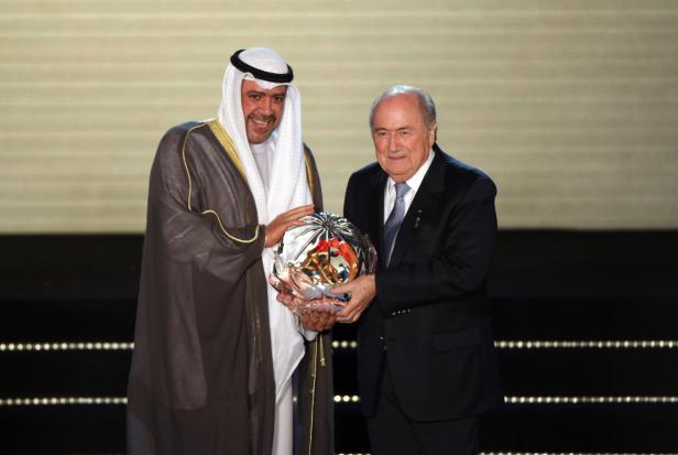 Ein Mann in traditioneller arabischer Kleidung und Sepp Blatter halten eine Trophäe.