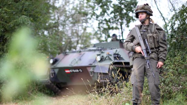 Eine Soldatin des österreichischen Bundesheeres steht mit einem Sturmgewehr vor einem Schützenpanzer.