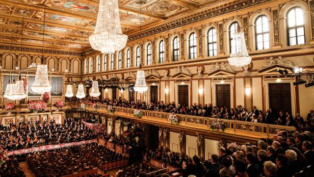 Ein Orchester spielt vor einem großen Publikum in einem opulenten Konzertsaal mit Kristalllüstern.