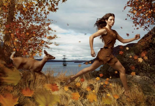 Eine Frau in einem braunen Kleid rennt mit einem Reh durch eine Herbstlandschaft mit einem Schiff im Hintergrund.