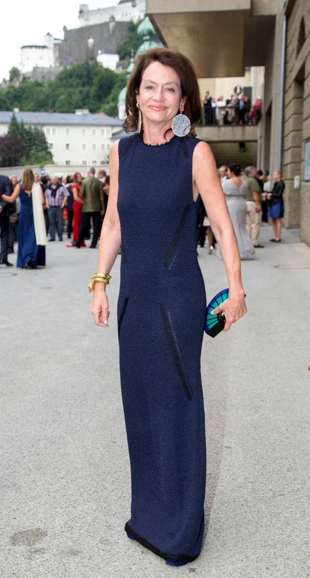 Eine Frau in einem blauen Kleid mit goldenem Armreif und Ohrringen vor der Festung Hohensalzburg.