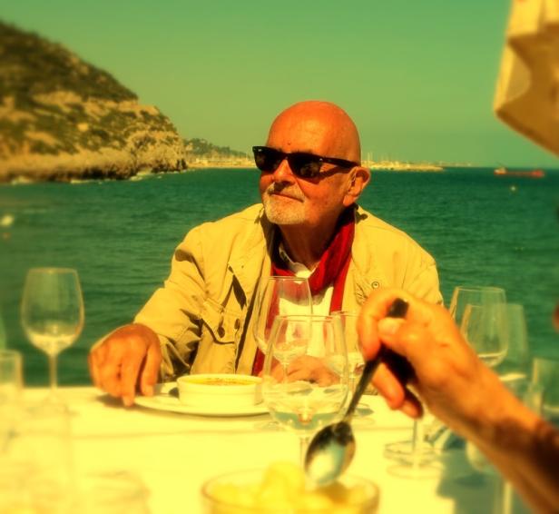 Ein älterer Mann mit Sonnenbrille sitzt an einem Tisch mit Blick auf das Meer.