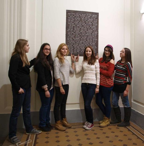 Sechs junge Frauen posieren vor einer Gedenktafel mit Namen an einer Wand.