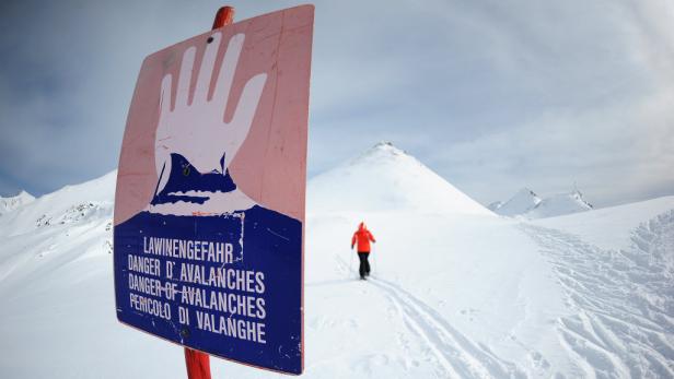Ein Lawinengefahr-Schild steht in einer verschneiten Berglandschaft, während eine Person im Hintergrund Ski fährt.