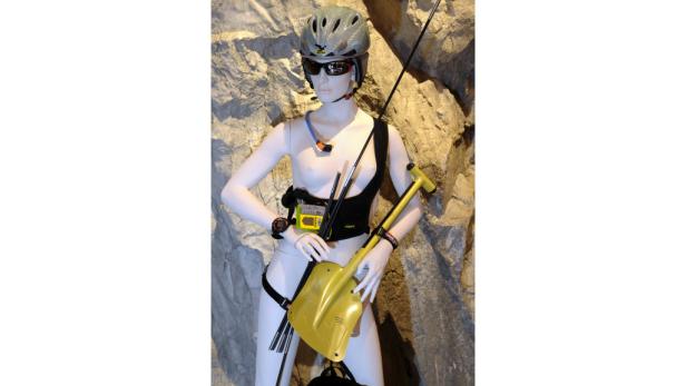 Eine Schaufensterpuppe mit Helm, Lawinenschutzausrüstung und einer gelben Schaufel vor einer Felswand.