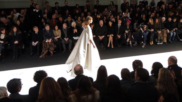 Eine Frau in einem langen, weißen Kleid läuft über einen Laufsteg vor einem Publikum.