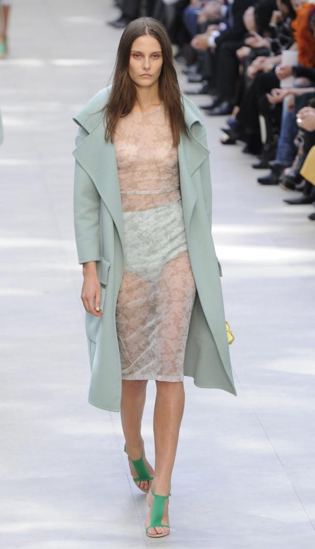 Eine Frau in einem transparenten Kleid und einem hellblauen Mantel auf einem Laufsteg.