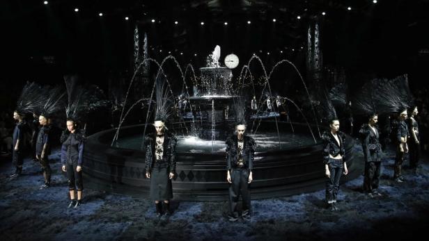 Models präsentieren dunkle Mode vor einem Brunnen mit einer Uhr im Hintergrund.