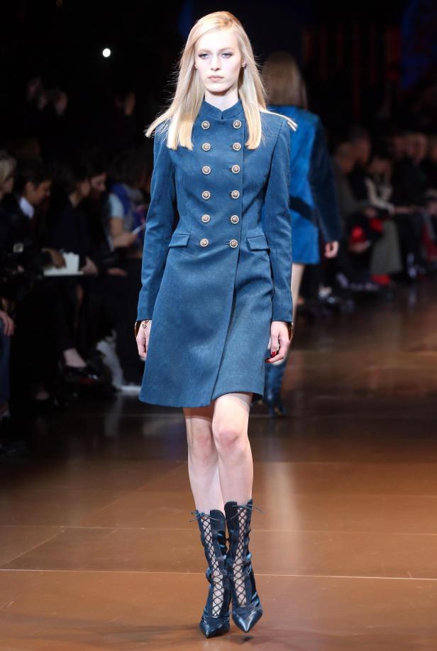 Eine blonde Frau präsentiert ein blaues Kleid mit goldenen Knöpfen und Schnürstiefeln auf dem Laufsteg.