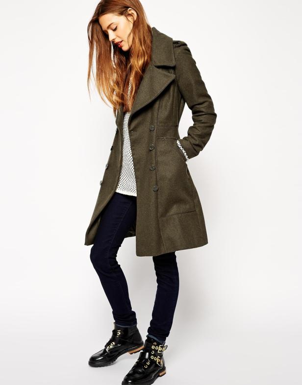 Eine Frau in einem olivgrünen Mantel, dunklen Jeans und schwarzen Stiefeln posiert.