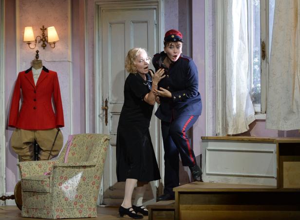 Zwei Frauen, eine in Zivil und eine in Uniform, erschrecken auf einer Theaterbühne.