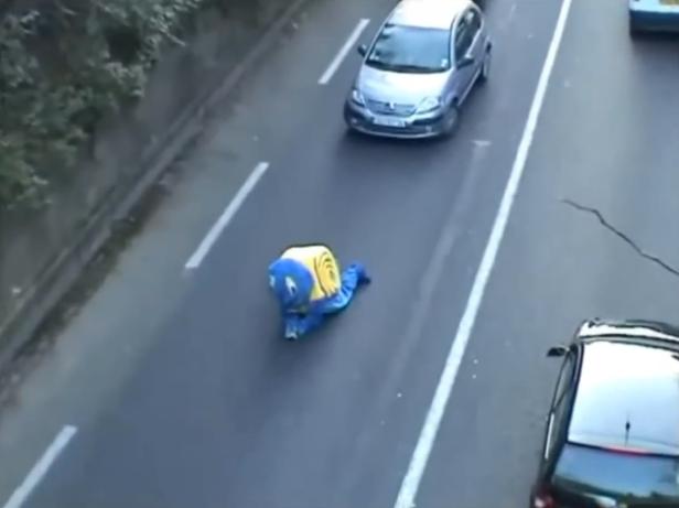 Eine Person im Schneckenkostüm kriecht auf einer Straße mit Autos.