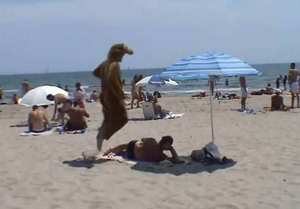 Eine Person im Kostüm springt über einen Mann, der am Strand liegt.