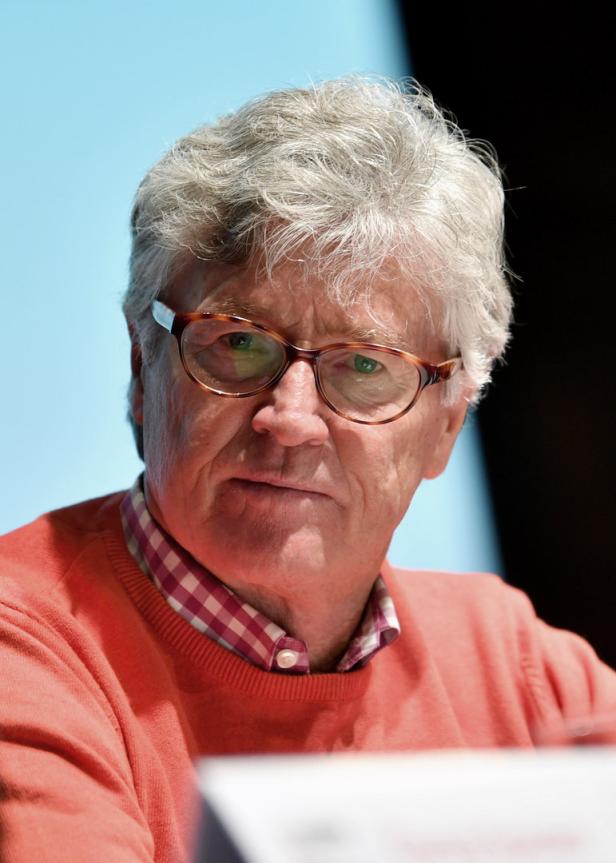 Ein Mann mit grauen Haaren und Brille trägt einen orangefarbenen Pullover.