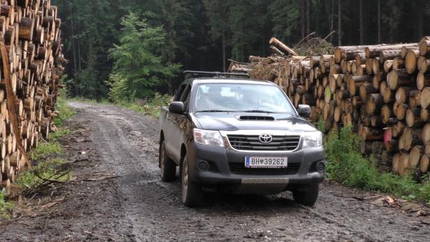Ein schwarzer Toyota Hilux fährt auf einer schlammigen Straße neben einem Holzstapel.