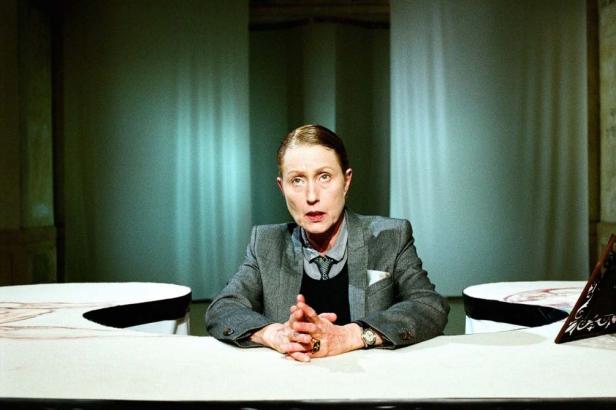 Eine Frau in einem grauen Blazer sitzt an einem Tisch und spricht.