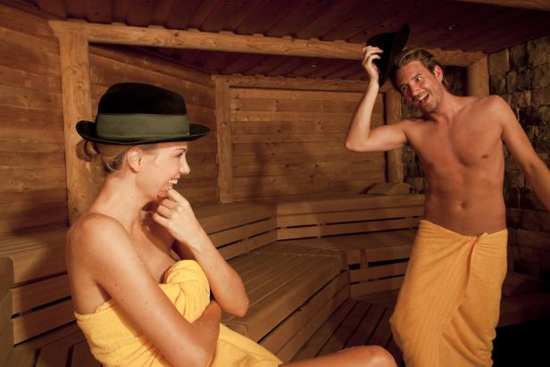 Eine Frau und ein Mann mit Hüten und Handtüchern in einer Sauna.