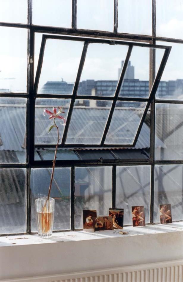 Ein offenes Fenster mit einer Lilie in einem Glas und Postkarten auf dem Fensterbrett.