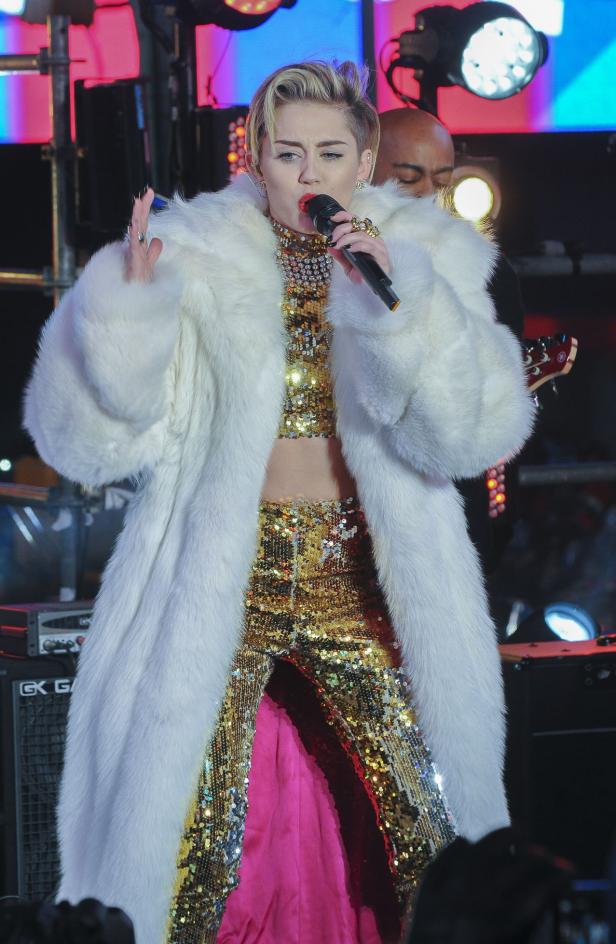 Miley Cyrus tritt in einem weißen Pelzmantel und einem goldenen Paillettenoutfit auf.
