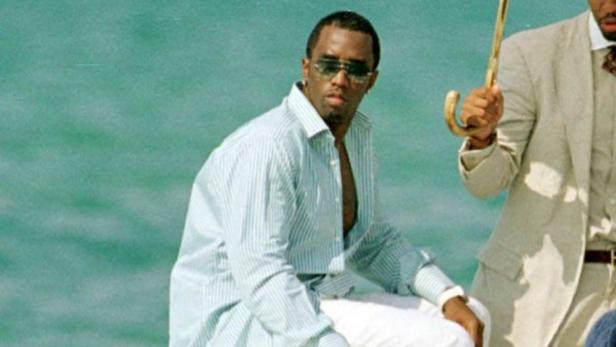 P. Diddy sitzt mit Sonnenbrille und offenem Hemd vor türkisblauem Wasser.