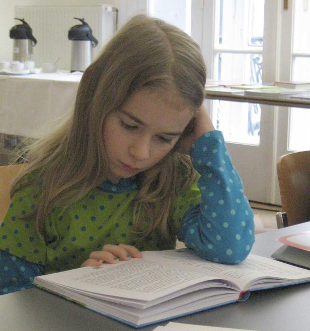 Ein Mädchen mit blonden Haaren liest ein Buch an einem Tisch.