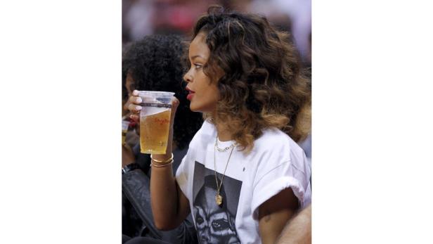 Rihanna mit lockigem Haar und T-Shirt hält einen Becher Bier in der Hand.