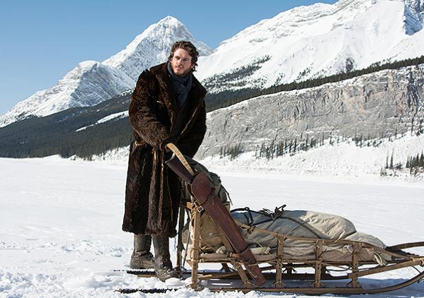 Ein Mann mit Pelzmantel steht mit einem Schlitten in einer verschneiten Berglandschaft.
