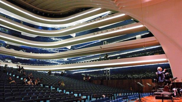 Ein Dirigent leitet ein Orchester in einem modernen Konzertsaal mit mehreren Rängen.