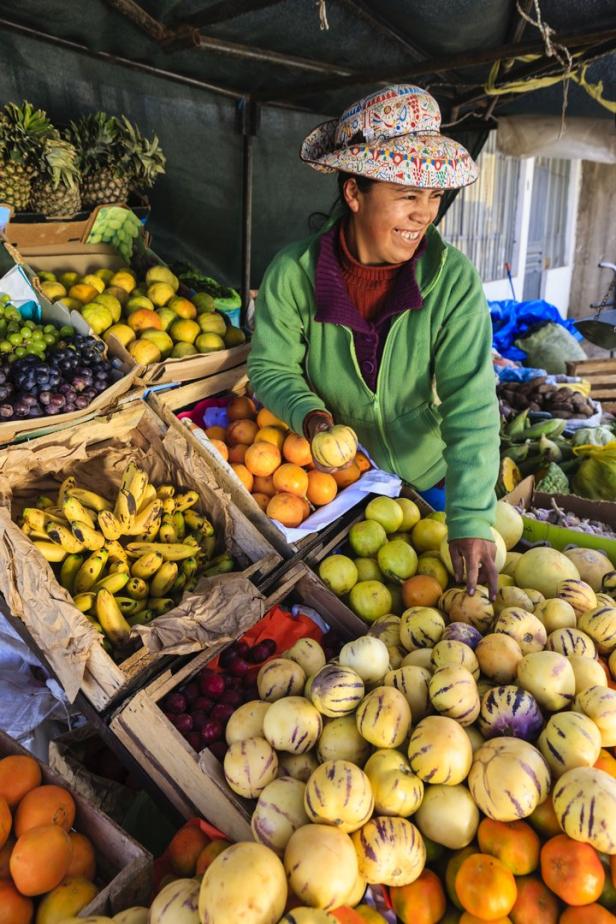Eine lächelnde Frau mit Hut verkauft Obst und Gemüse auf einem Markt.