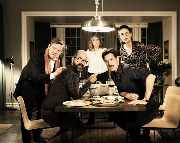 Fünf Personen sitzen oder stehen an einem Esstisch und blicken überrascht in die Kamera.