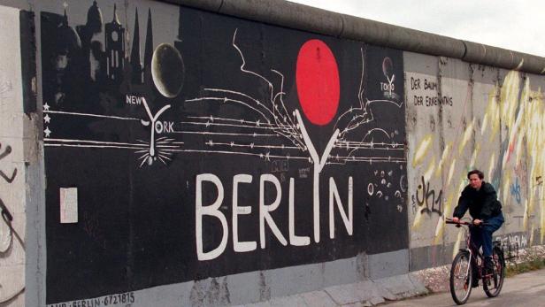 Ein Mann fährt mit dem Fahrrad vor einem bemalten Abschnitt der Berliner Mauer.
