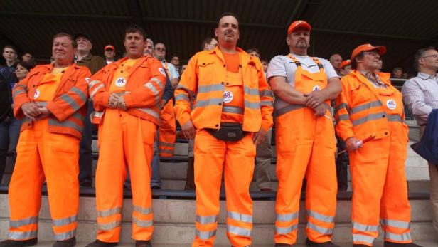 Eine Gruppe Männer in orangefarbenen Overalls steht auf einer Tribüne.