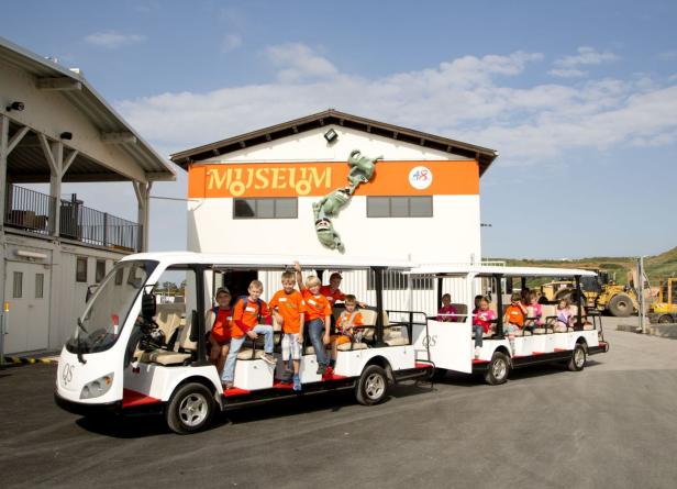 Eine Gruppe Kinder fährt mit einem Zug vor einem Museum.