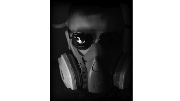 Eine Person mit Schutzmaske und Sonnenbrille, in deren Gläsern sich ein Feuer spiegelt.