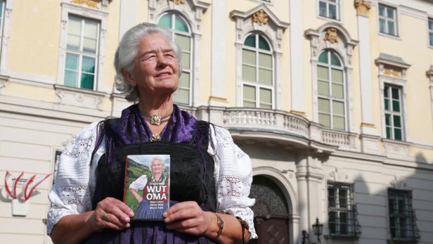 Eine ältere Frau im Dirndl hält ihr Buch „Wut Oma“ vor einem Gebäude.