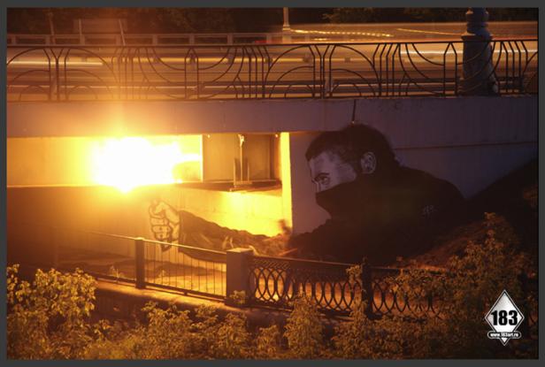 Ein Wandgemälde unter einer Brücke zeigt eine Person mit bedecktem Gesicht und erhobener Faust.