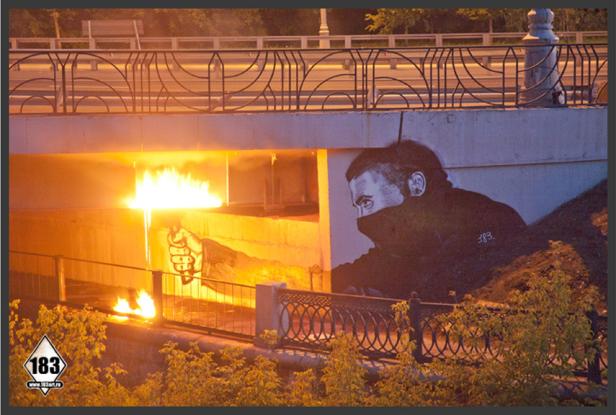 Unter einer Brücke brennt ein Feuer neben einem Graffiti eines Mannes mit Maske.