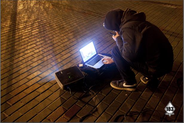 Eine Person hockt im Freien vor einem Laptop und einem Projektor.