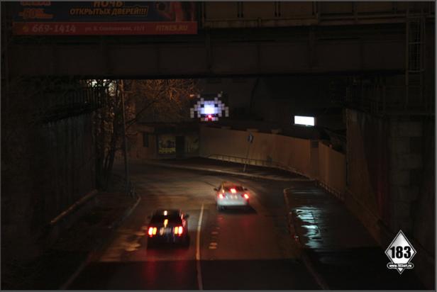 Zwei Autos fahren nachts unter einer Brücke hindurch, über der ein Space Invader-Motiv leuchtet.