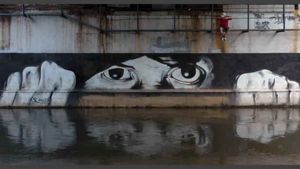 Ein großes Wandgemälde mit Augen und Händen, das sich im Wasser spiegelt; eine Person sitzt darüber.